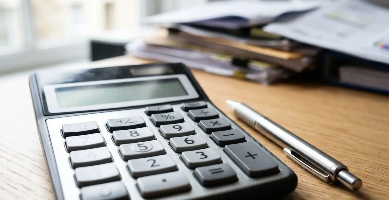 Vue rapprochée d'une calculatrice contemporaine posée sur des documents administratifs floutés avec un stylo métallique, éclairés par une lumière naturelle latérale